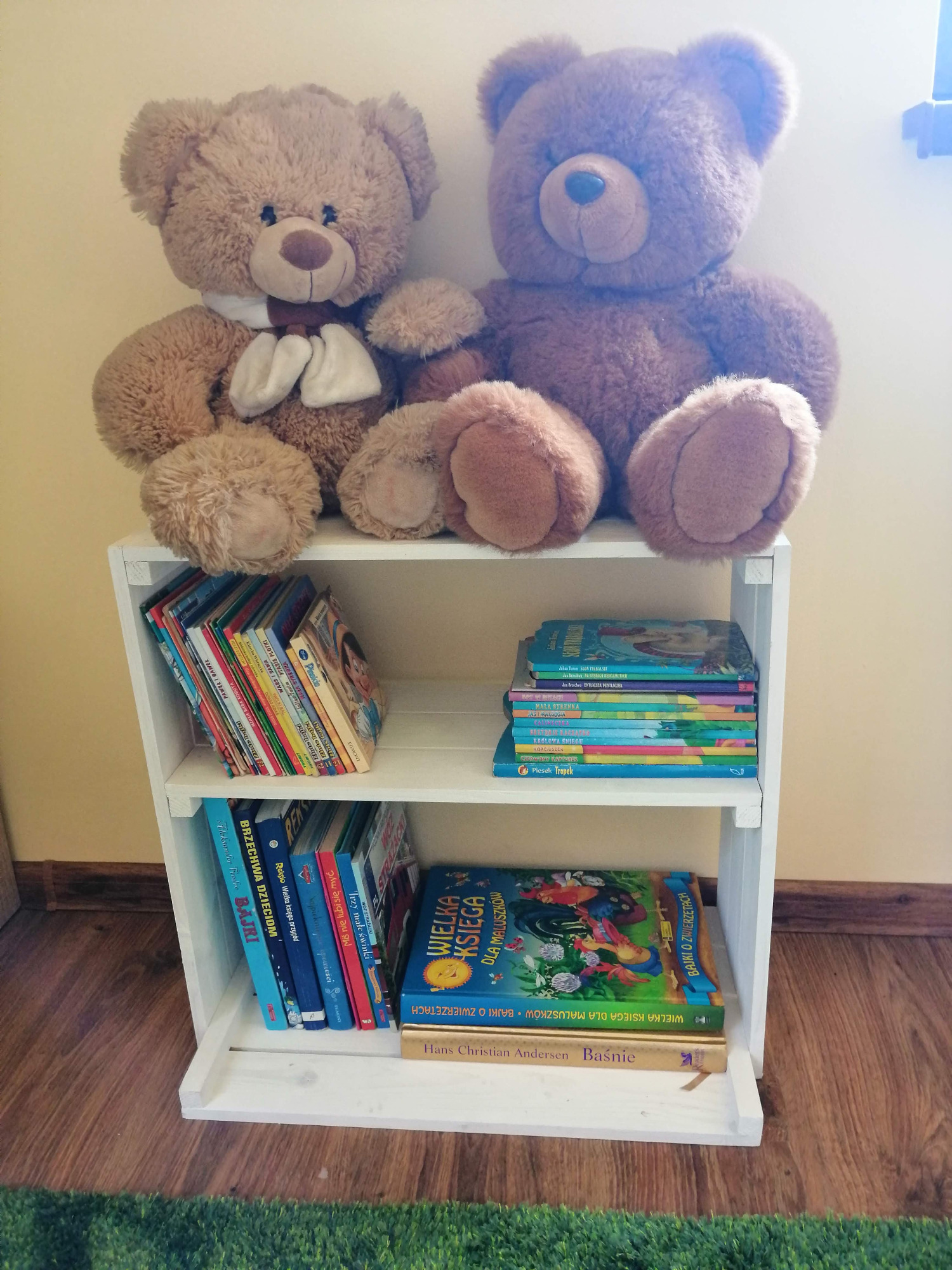 Biały, drewniany regał z trzema półkami, na których ułożone są książki dla dzieci i dwa pluszowe misie na górze, ustawiony na podłodze wyłożonej panelami i sztuczną trawą.