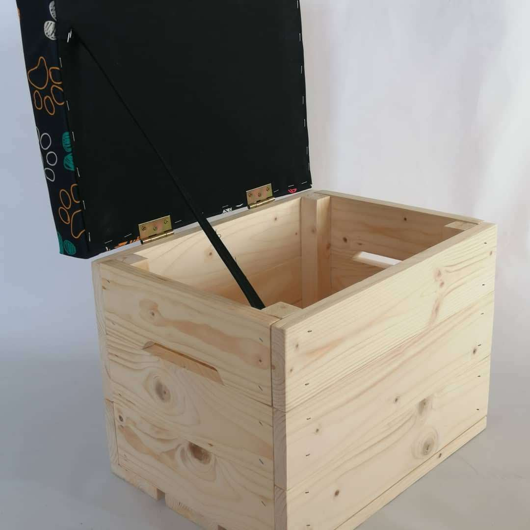 Otwarty, surowy drewniany kufer z podnoszonym wiekiem obitym czarną tkaniną w kolorowe wzory, widoczne zawiasy i mechanizm podtrzymujący wieko.