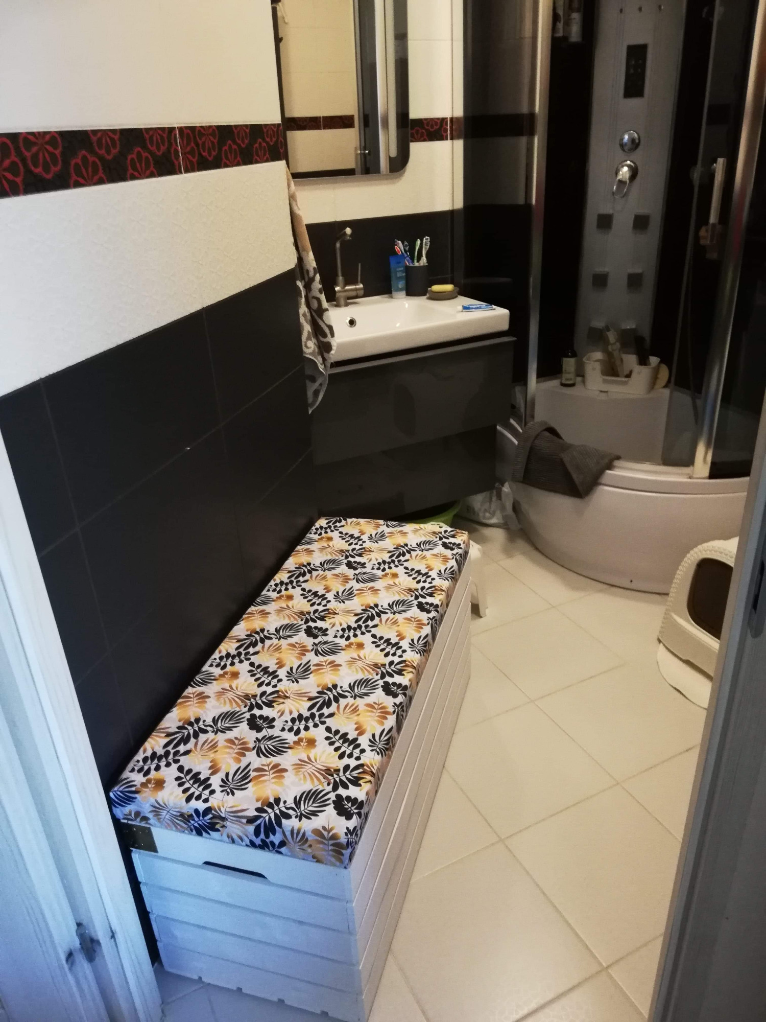 Biała skrzynia-pufa z wodoodpornym obiciem w liście, umieszczona w łazience z czarno-białymi płytkami, obok kabiny prysznicowej i umywalki.