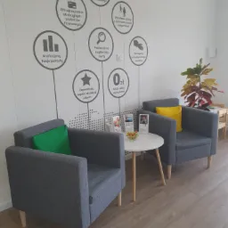 Poczekalnia biura z dwoma szarymi fotelami z poduszkami w kolorze limonkowym i żółtym, stolikiem kawowym z miseczką owoców i ulotkami, oraz ścianą z infografiką o ofercie firmy.