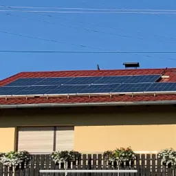 Dwa panele słoneczne na czerwonym dachu domu jednorodzinnego z ceglanym kominem, widok z dołu, w tle błękitne niebo i linie energetyczne.