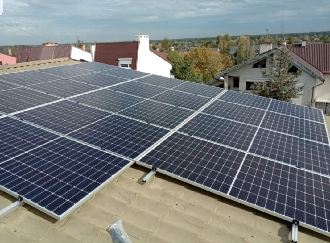 Panel słoneczny na dachu domu jednorodzinnego, widoczne mocowania i fragment narzędzia w dolnym rogu, w tle zabudowa mieszkalna i drzewa.