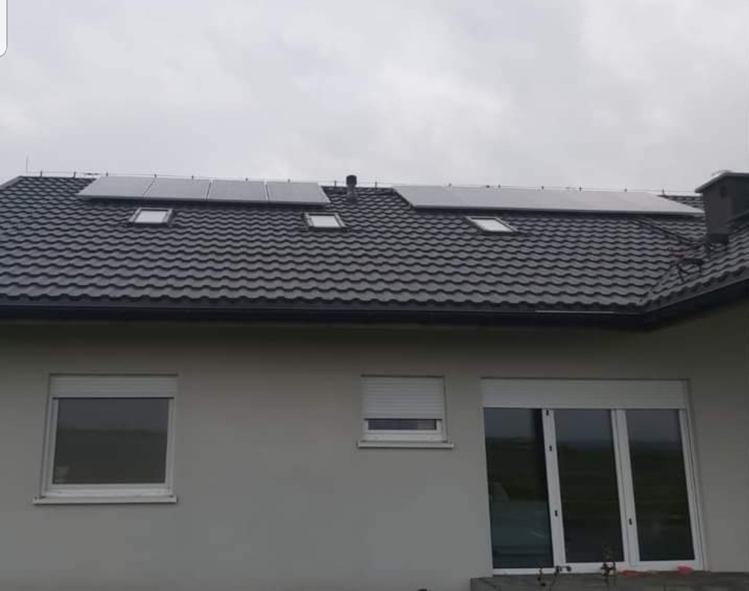Panele słoneczne na dachu domu jednorodzinnego z szarą dachówką i oknami z roletami zewnętrznymi, pochmurne niebo w tle.