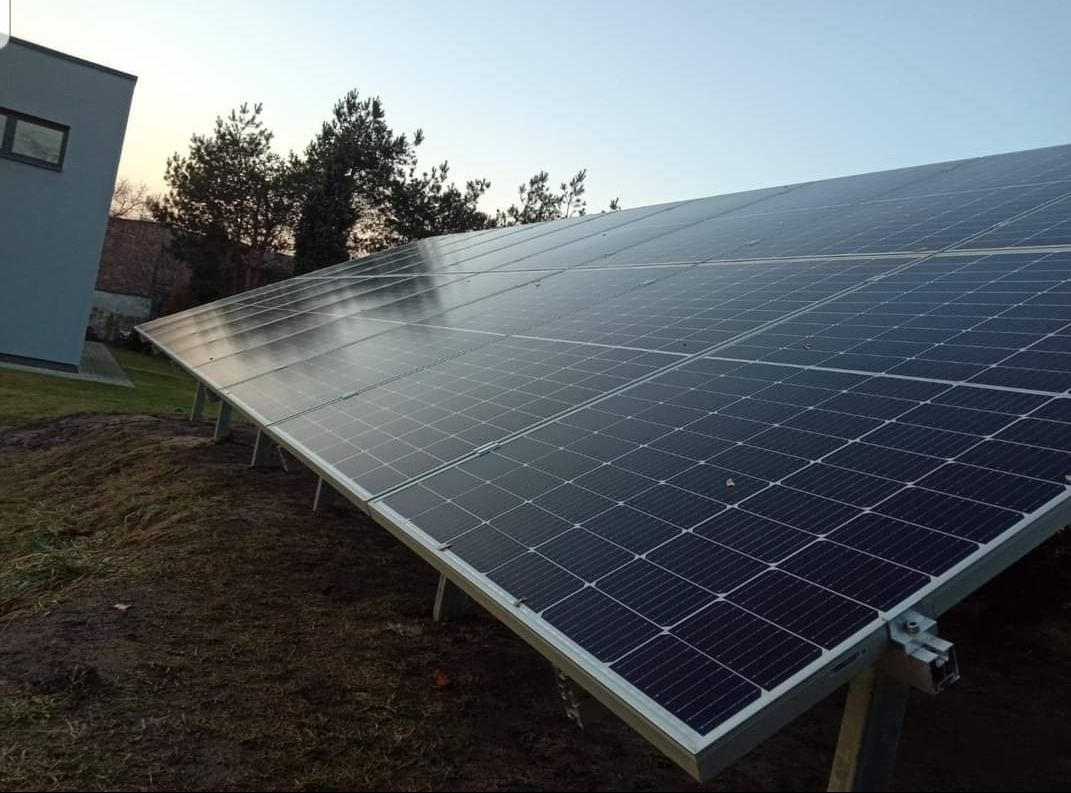 Instalacja paneli słonecznych na gruncie, widoczne metalowe podpory, w tle budynek mieszkalny i drzewa, pochmurne niebo.