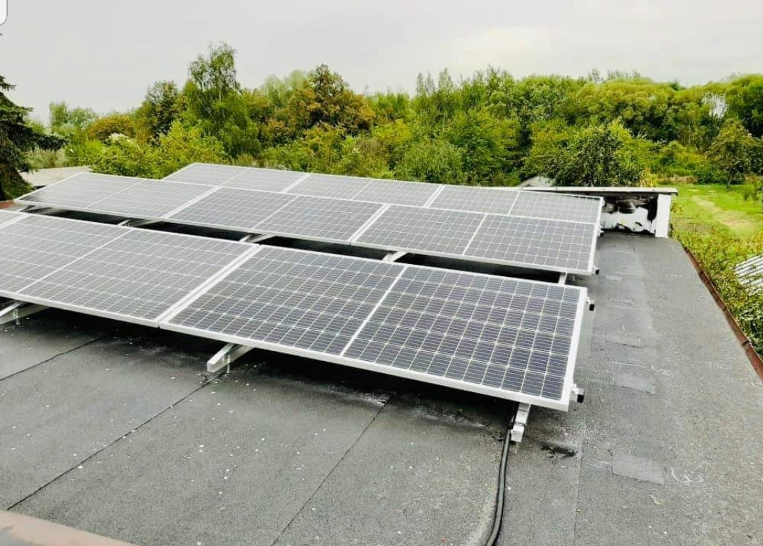 Panel fotowoltaiczny zamontowany na płaskim dachu pokrytym papą, z widocznymi przewodami i zielonym tłem drzew.