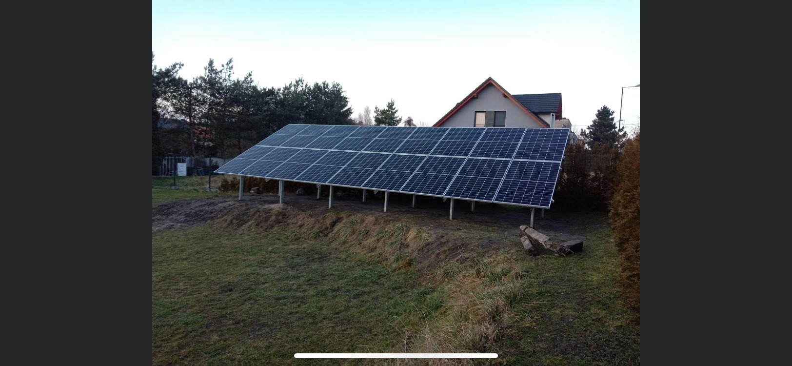 Instalacja paneli słonecznych na metalowej konstrukcji w ogrodzie, w tle dom jednorodzinny i drzewa.