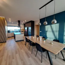 Nowoczesna kuchnia z jadalnią: drewniana podłoga, stół z czarnymi krzesłami, wyspa kuchenna z drewnianymi frontami, niebieska ściana z odbiciem roślinności, designerskie lampy wiszące.