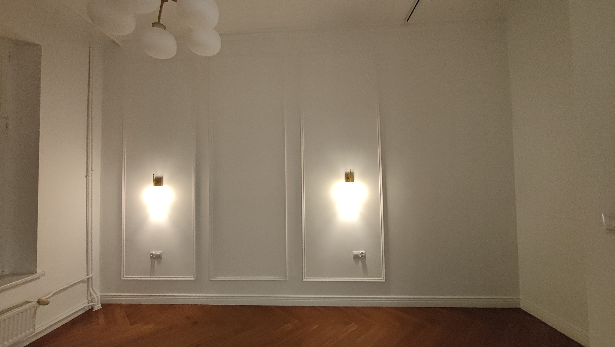 Puste, białe wnętrze z parkietem, dekoracyjnymi panelami ściennymi, dwoma identycznymi kinkietami z ciepłym światłem i widoczną instalacją elektryczną z gniazdkami.