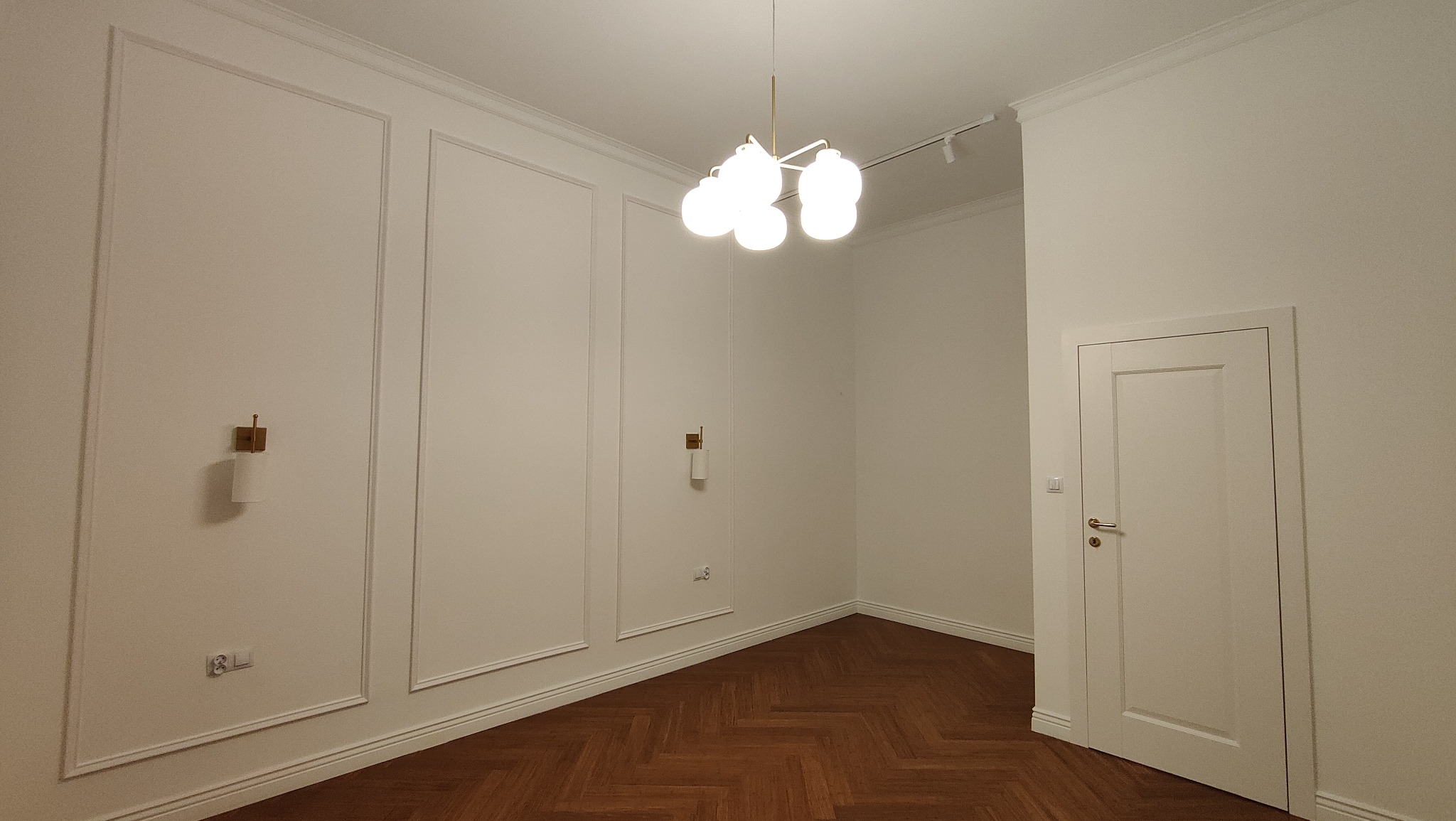 Puste, białe wnętrze z parkietem w jodełkę, ozdobnymi listwami ściennymi, nowoczesną lampą sufitową i kinkietami z białymi abażurami.