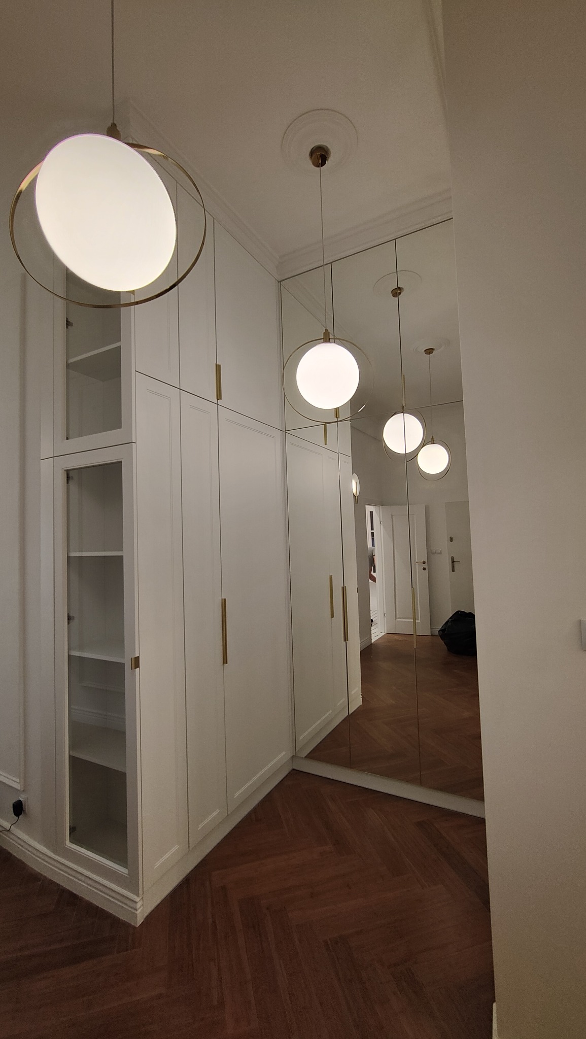 Biała, zabudowana szafa z przeszklonymi witrynami, złotymi uchwytami i lustrzanymi drzwiami, oświetlona wiszącymi, okrągłymi lampami z mosiężnymi obręczami, na podłodze parkiet ułożony w jodełkę.