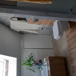 Nowoczesna łazienka z drewnopodobnymi płytkami na podłodze i ścianie, białą armaturą i szafkami oraz rośliną doniczkową na blacie umywalki.