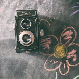 Spółka Rzeszowska - Czarno-biały aparat dwuobiektywowy marki Start zawieszony na sznurku na tle tablicy kredowej z rysunkiem kwiatka.