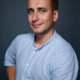 Portret uśmiechniętego mężczyzny z niebieskimi oczami i blond włosami w błękitnej koszuli na ciemnoszarym tle.