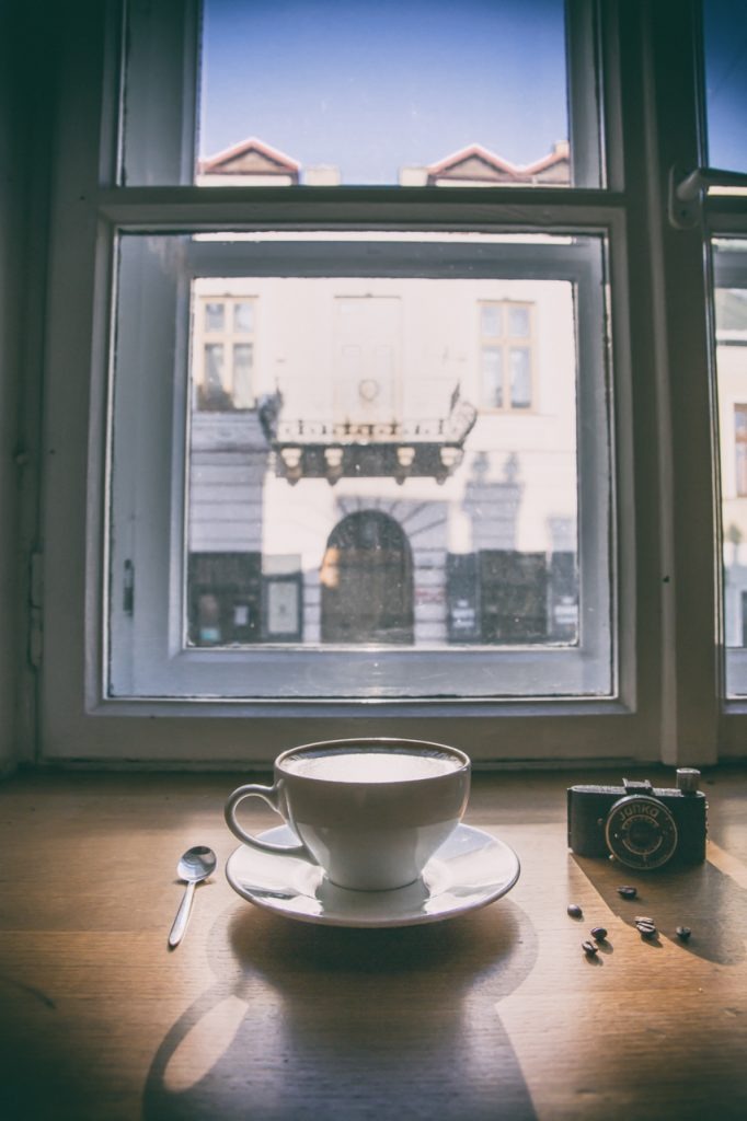 Filiżanka kawy na drewnianym parapecie z widokiem na budynek przez okno, obok leży srebrna łyżeczka, miniaturowy aparat fotograficzny i kilka ziaren kawy.