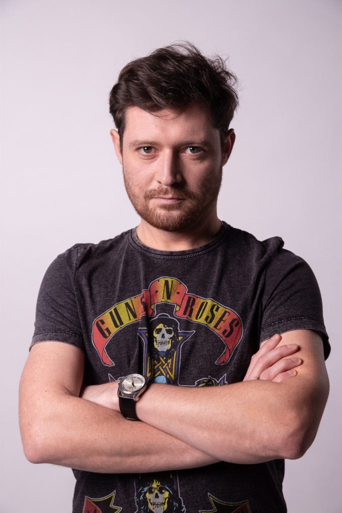 Portret mężczyzny z brodą w koszulce Guns N' Roses, z zegarkiem na nadgarstku i skrzyżowanymi rękami na białym tle.