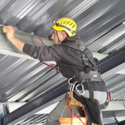 Pracownik w uprzęży, z żółtym kaskiem i lampką czołową, czyści metalową konstrukcję sufitu gąbką, widoczne elementy konstrukcyjne i wiadro.