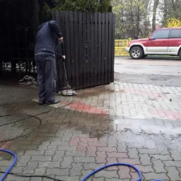 Czyszczenie kostki brukowej przed posesją za pomocą myjki ciśnieniowej; widoczna różnica między umytą a brudną powierzchnią, drewniana brama i czerwono-biały samochód w tle.
