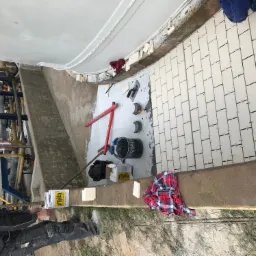 Instalacja białych płytek na ścianie w obiekcie przemysłowym, widoczny pracownik w niebieskim kasku, narzędzia glazurnicze, poziomica, wiadro z zaprawą i resztki materiałów budowlanych.