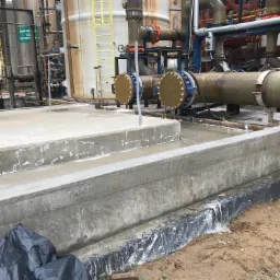 Betonowa konstrukcja zbiornika z wystającymi rurami, widoczne ślady po zalaniu, zabezpieczenie folią od strony gruntu.