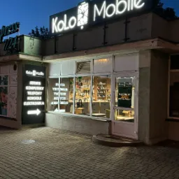 Fasada serwisu telefonów Kolo Mobile oświetlona neonowym szyldem i bocznym panelem z listą usług, widziana z poziomu brukowanego chodnika o zmierzchu.