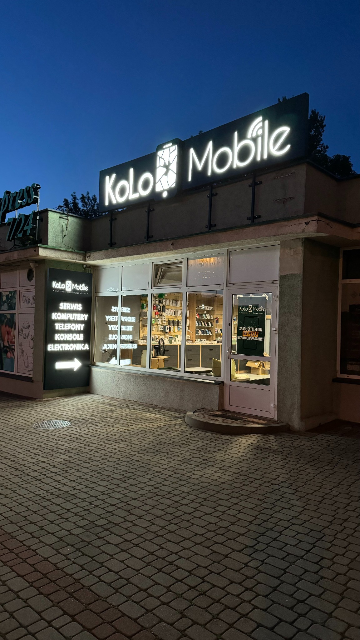 Fasada serwisu telefonów Kolo Mobile oświetlona neonowym szyldem i bocznym panelem z listą usług, widziana z poziomu brukowanego chodnika o zmierzchu.