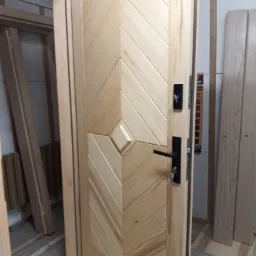 Otwarta, surowa drewniana brama z ukośnym wzorem desek i czarnym zamkiem, widoczne klucze w zamku, w tle fragmenty ościeżnicy i desek.