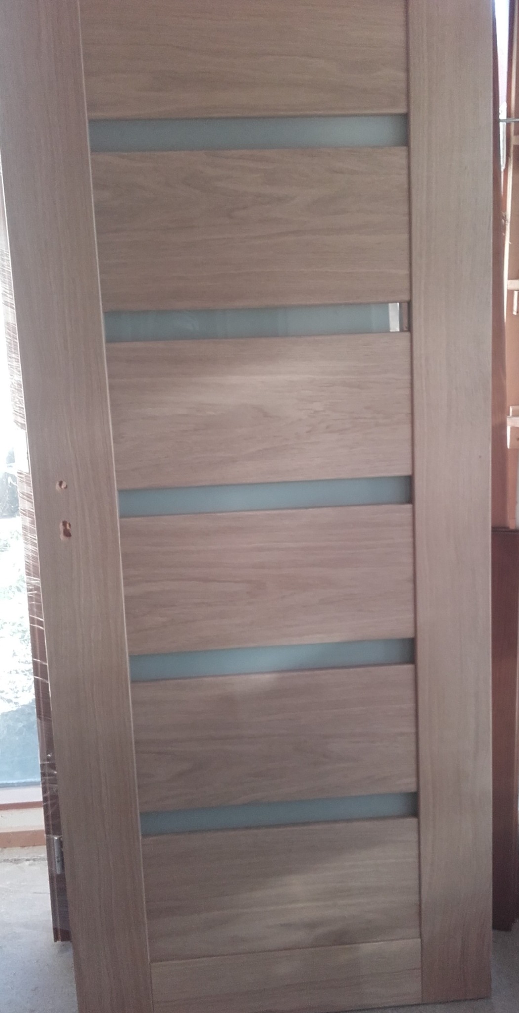 Niedokończona montażowo, drewniana konstrukcja z czterema poziomymi przeszkleniami, widoczne otwory na klamkę, częściowo owinięta folią ochronną.