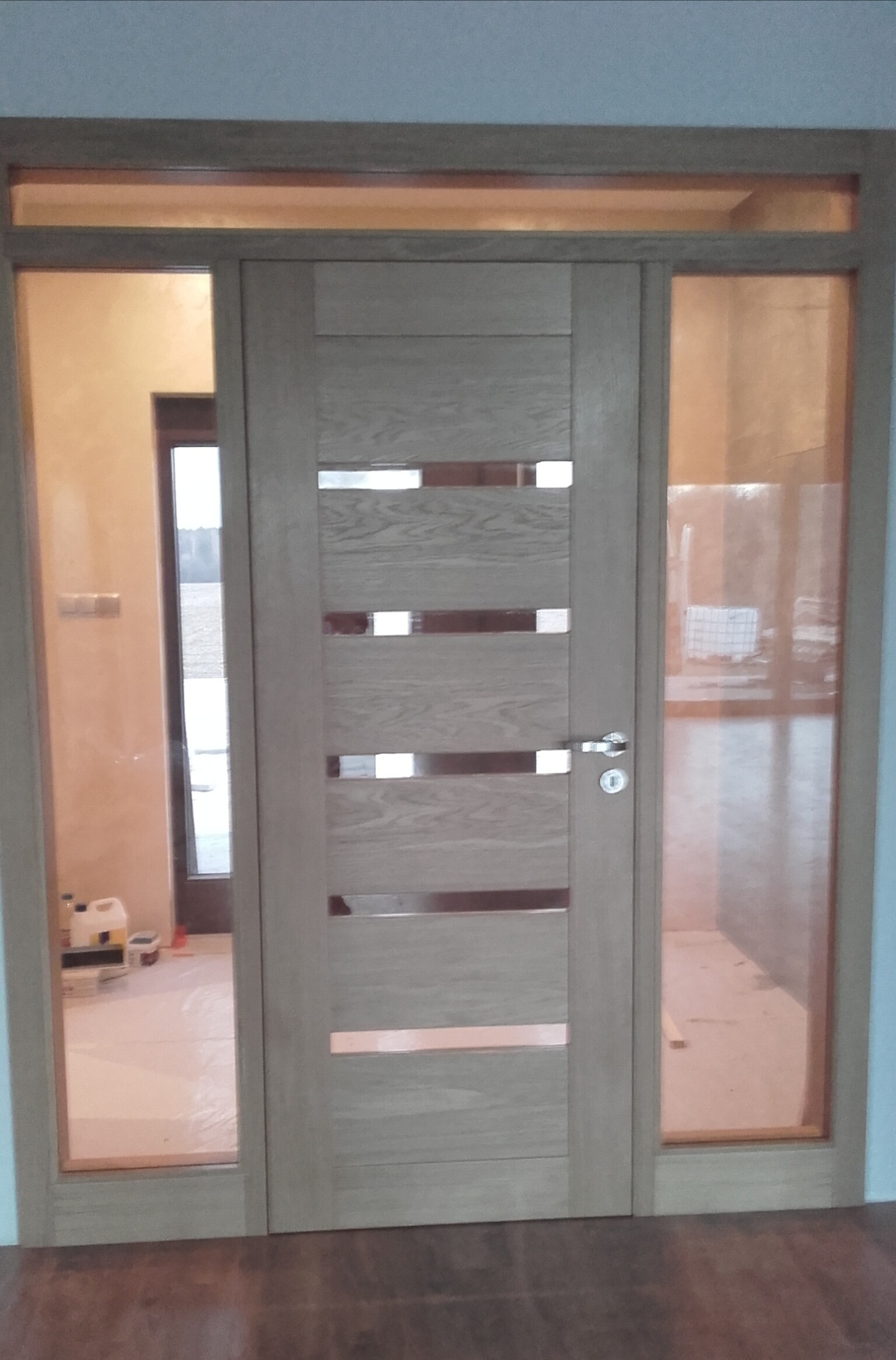 Jasne drzwi wewnętrzne z przeszkleniami wstawione w ościeżnicę z bocznymi naświetlami, widoczne przygotowania do malowania pomieszczenia w tle.