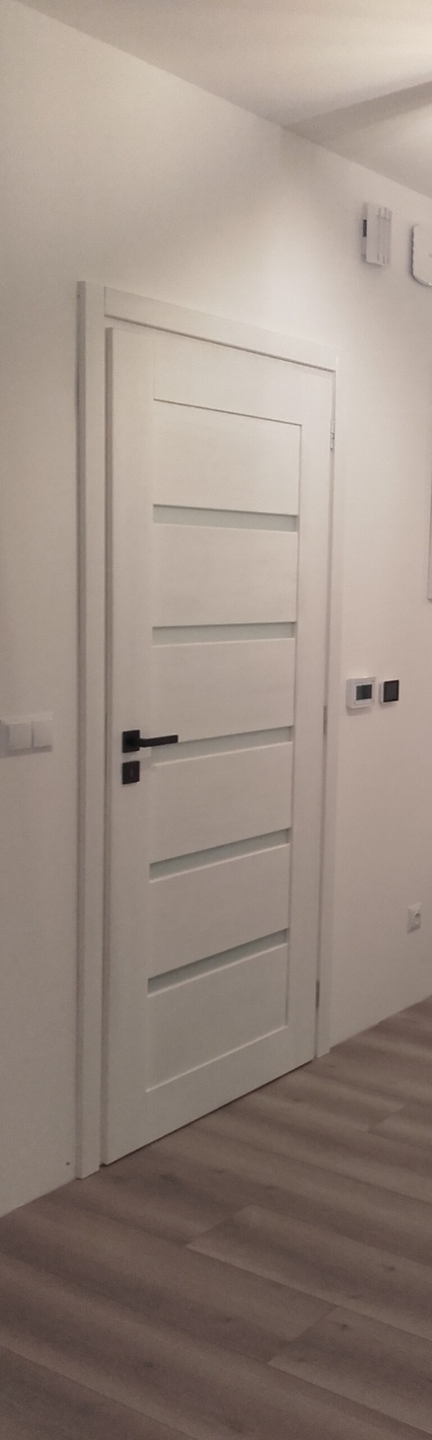 Białe drzwi wewnętrzne z poziomymi wgłębieniami i czarną klamką na tle białej ściany i drewnopodobnej podłogi.