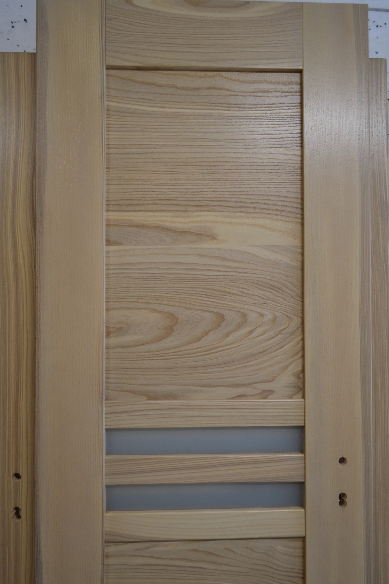 Jasne drewniane drzwi z widocznym usłojeniem, z matowymi, szklanymi wstawkami w dolnej części i otworami na klamkę, prezentowane w pionowym kadrze.