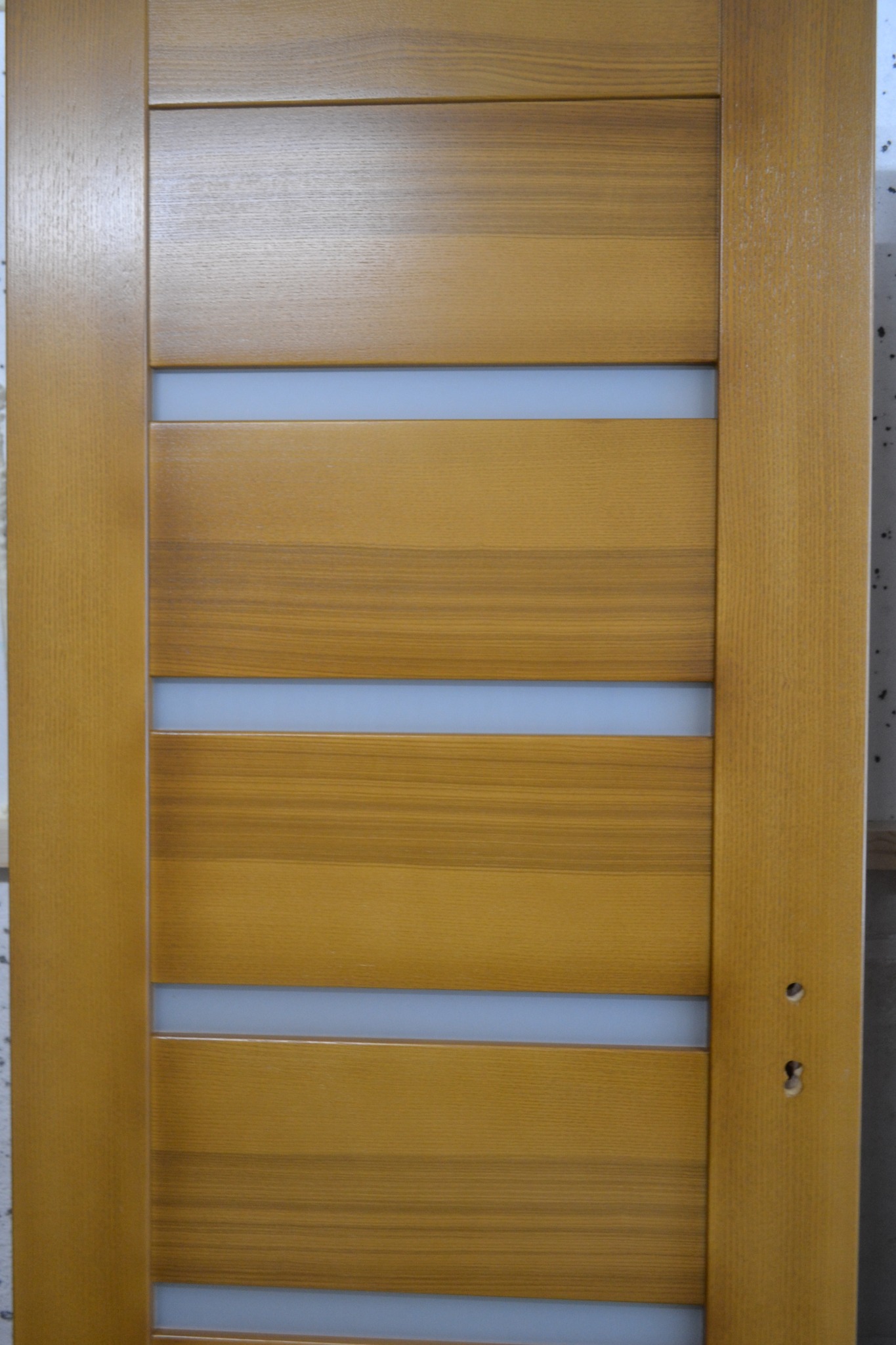 Drewniane skrzydło drzwiowe w odcieniach brązu, z poziomymi wstawkami z mlecznego szkła, widoczne otwory pod klamkę i zamek.