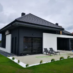 Nowoczesny dom z grafitową elewacją, białymi akcentami i roletami zewnętrznymi, widok z ogrodu, dwa leżaki na tarasie.