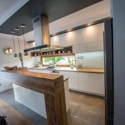 Nowoczesna kuchnia z białymi szafkami, drewnianymi blatami i wyspą z blatem z drewna, oświetlenie punktowe i wiszące lampy, widok na okno z zielenią.