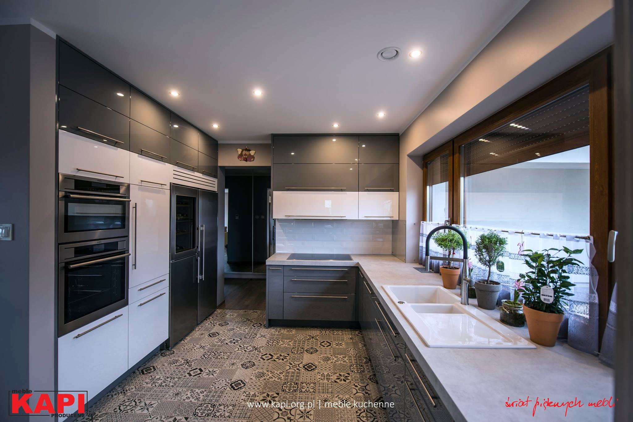 Nowoczesna kuchnia z szarymi i białymi szafkami na wysoki połysk, wzorzystą podłogą i roślinami w doniczkach przy zlewie.