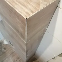 Narożnik obudowany płytkami imitującymi drewno, z widocznymi spoinami i wiaderkiem z zaprawą w dolnej części kadru.