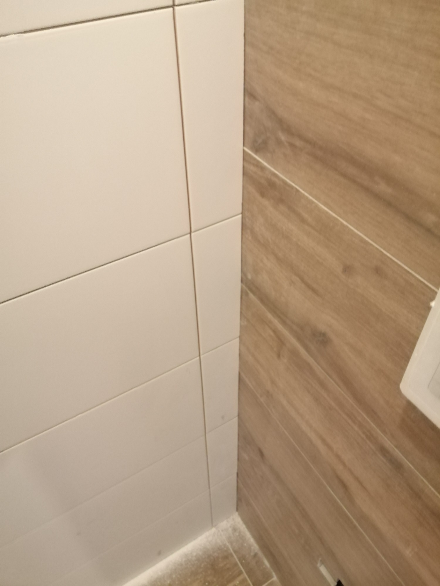 Połączenie białych, prostokątnych płytek ceramicznych z płytkami imitującymi drewno na ścianie, widoczne minimalne ślady fugi na podłodze.