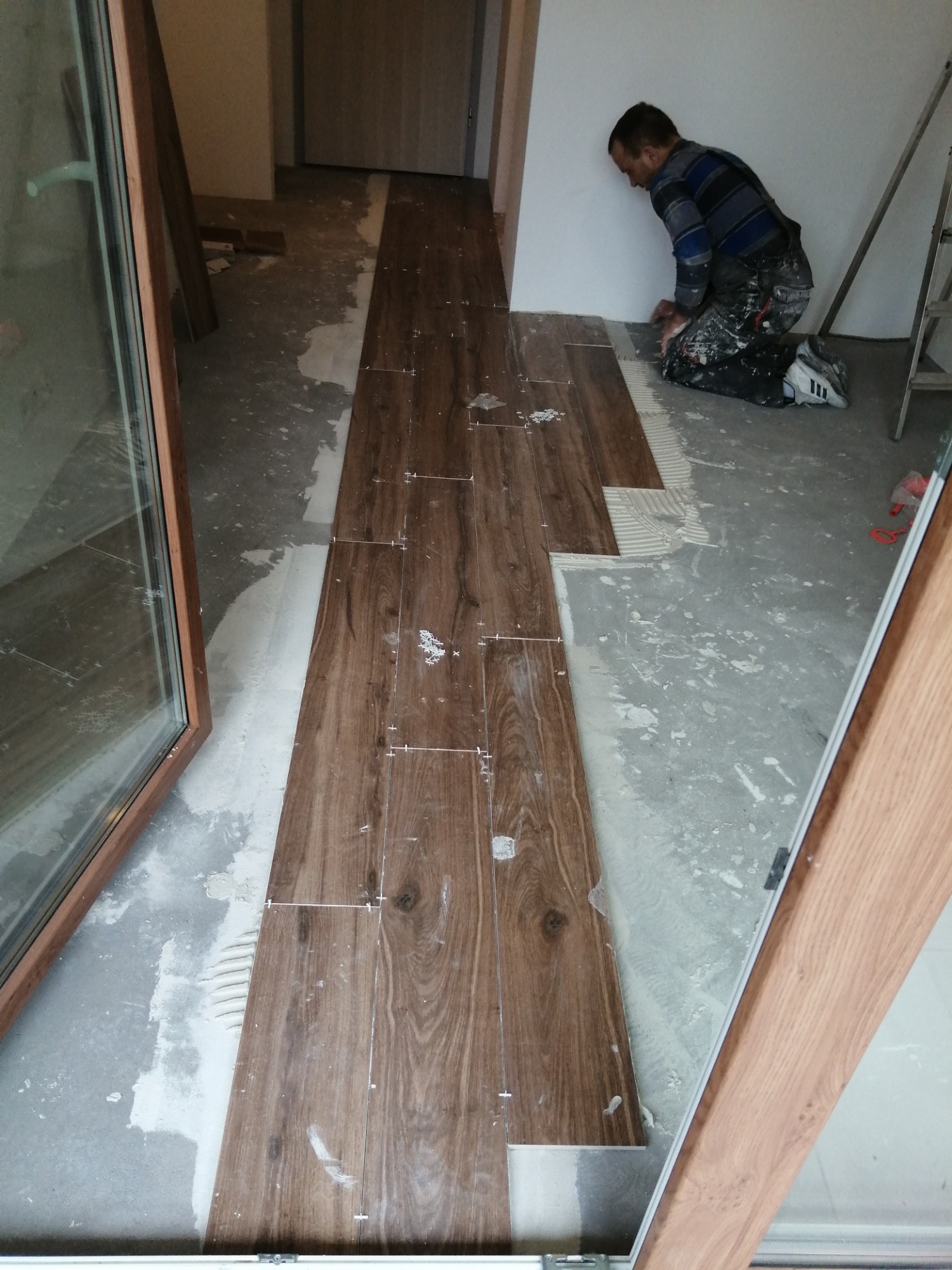 Układanie podłogi z drewnopodobnych płytek: glazurnik na kolanach dociska płytkę do podłoża, widoczna fuga i krzyżyki dystansowe, częściowo ułożona podłoga w korytarzu.