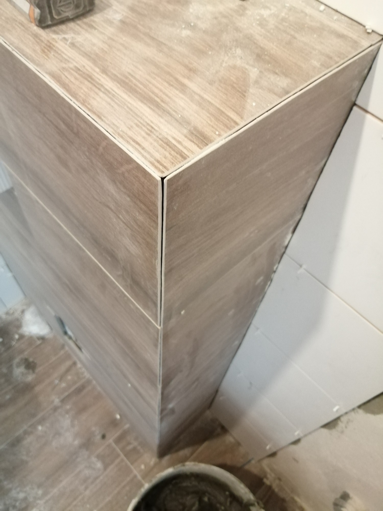 Narożnik obudowany płytkami imitującymi drewno, z widocznymi spoinami i wiaderkiem z zaprawą w dolnej części kadru.