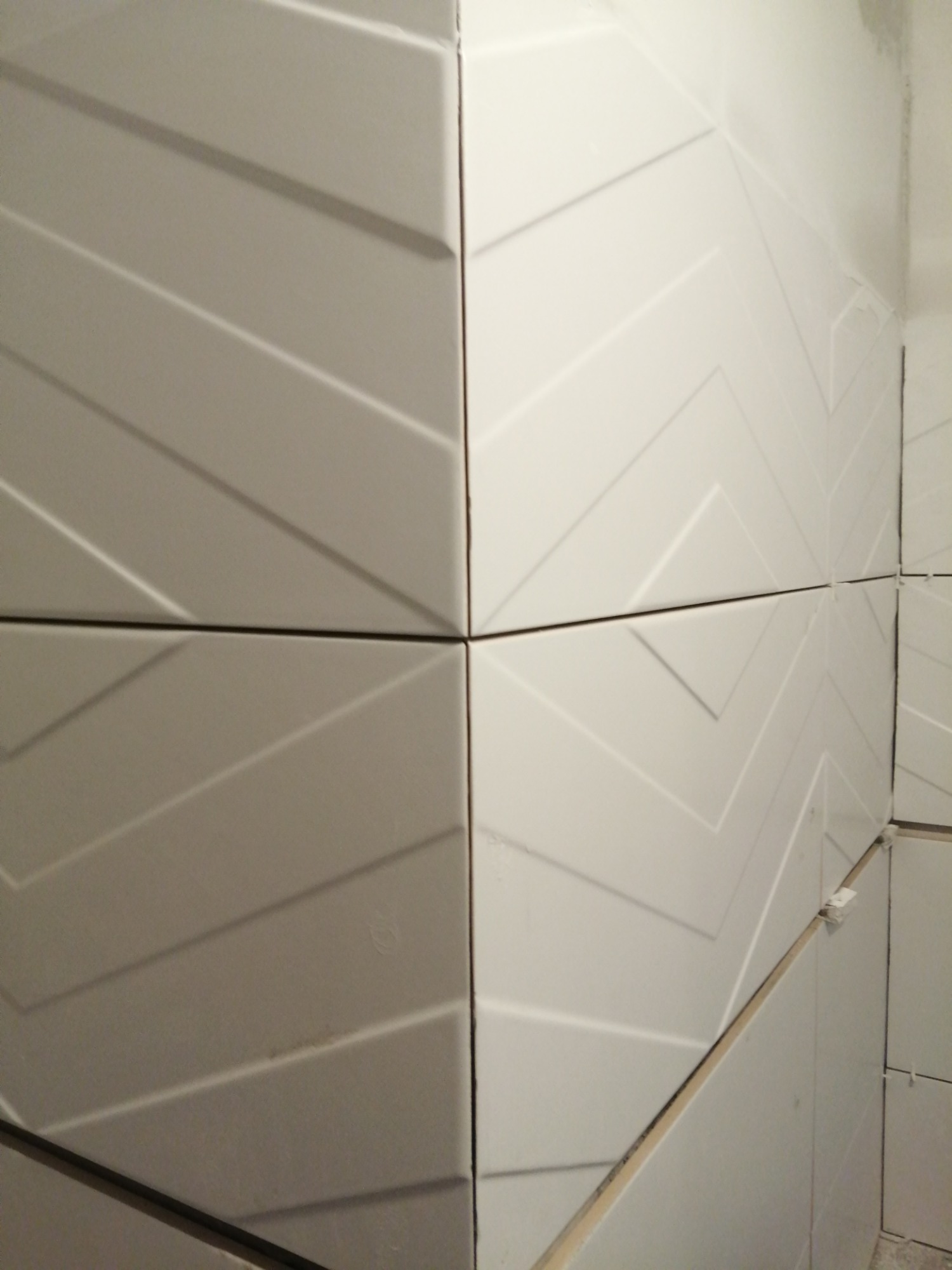 Ułożone białe płytki ścienne z trójwymiarowym wzorem w jodełkę, widoczne plastikowe krzyżyki dystansowe w spoinach.