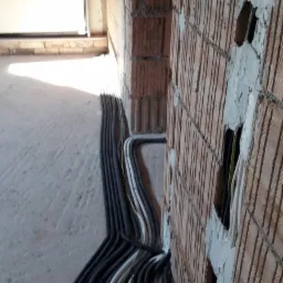 Ściana z czerwonej cegły z wyciętymi otworami, w których widać kable, a na podłodze leżą rury ochronne z kablami. Widoczny surowy beton i brama garażowa w tle.