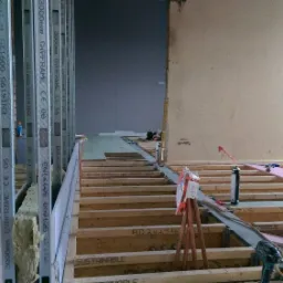 Wnętrze w trakcie remontu: widoczne metalowe profile konstrukcyjne z wypełnieniem z wełny mineralnej, drewniany szkielet podłogi, narzędzia, puszka z pianką montażową i biały kask budowlany...