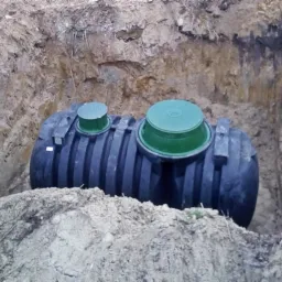 Czarny zbiornik na szambo z zielonymi pokrywami umieszczony w wykopie ziemnym, widoczna rura kanalizacyjna po lewej stronie.