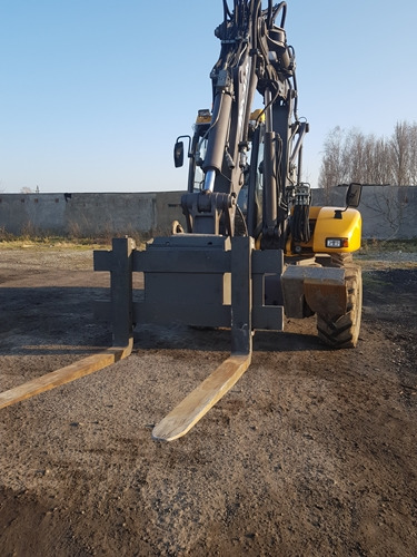 Żółta koparka kołowa Volvo z zamontowanymi widłami do palet stoi na ubitym gruncie, w tle betonowy mur i drzewa bez liści, widok z przodu na widły.