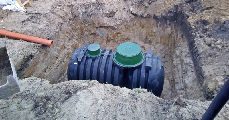 Czarny zbiornik na szambo z zielonymi pokrywami umieszczony w wykopie ziemnym, widoczna rura kanalizacyjna po lewej stronie.