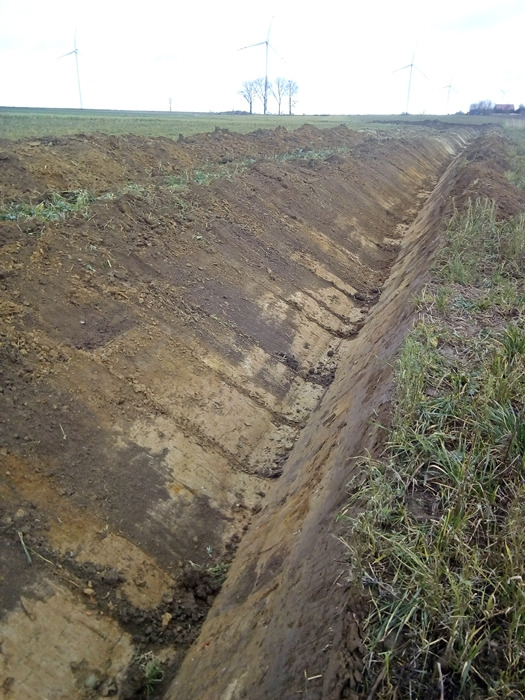 Długi, prosty rów wykopany w polu, z widocznymi śladami gąsienic maszyny na ścianach, w tle turbiny wiatrowe i drzewa.