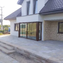 Ułożony taras z kostki brukowej w odcieniach beżu i szarości, prowadzący do nowoczesnego domu z dużymi przesuwnymi drzwiami tarasowymi, widoczne niedokończone obrzeża i ziemia.