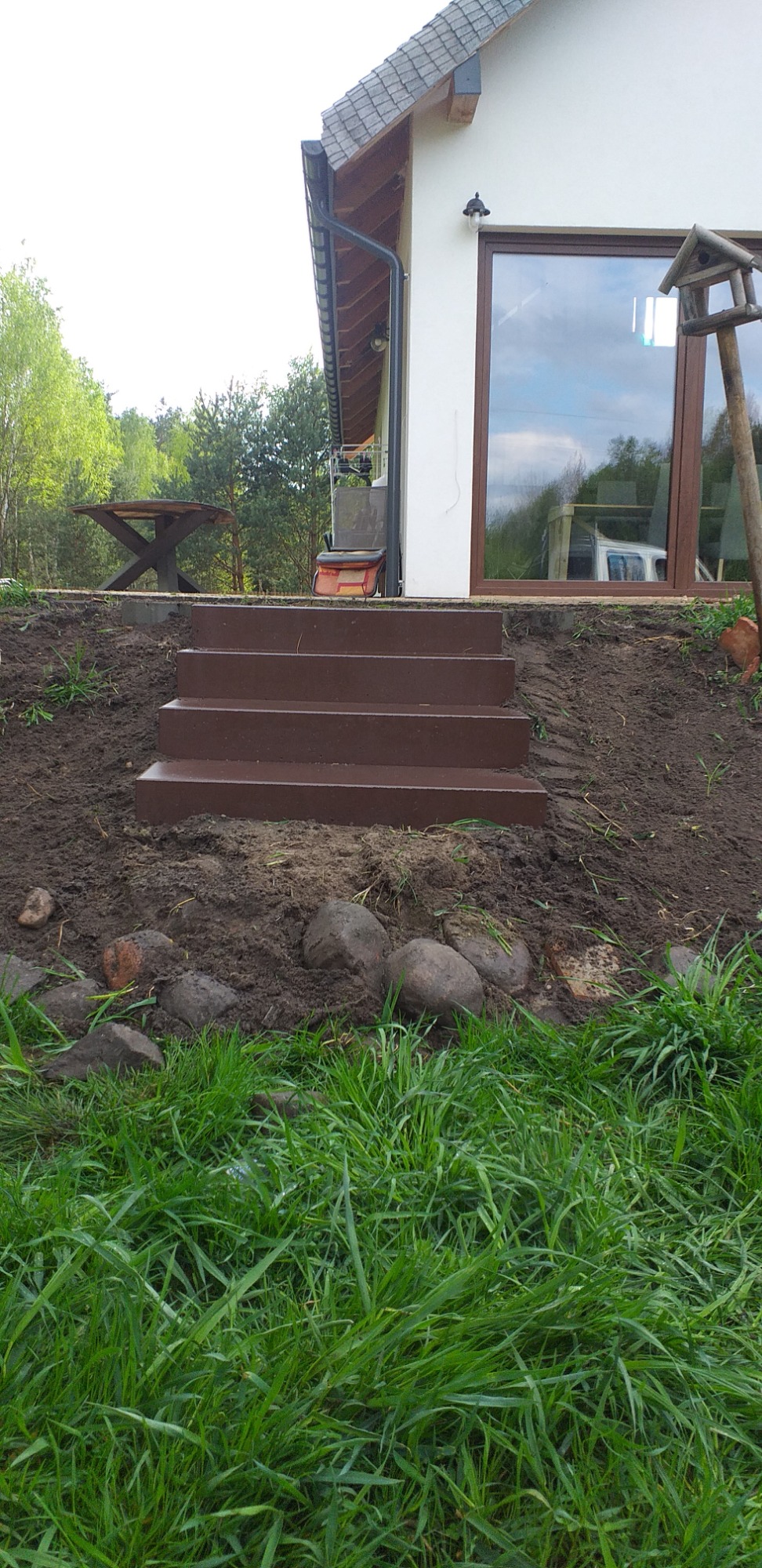 Nowe, brązowe schody z kostki brukowej prowadzące do domu z białymi ścianami i brązowymi ramami okiennymi, widoczne kamienie dekoracyjne u podstawy schodów i bujna trawa z przodu.