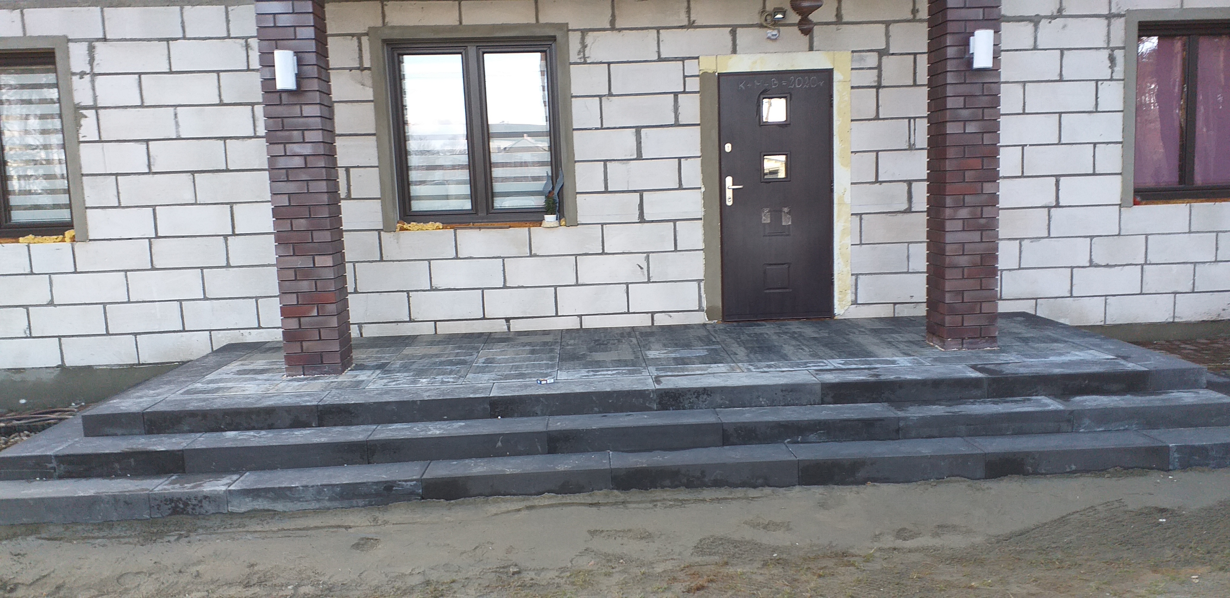 Schody wejściowe wyłożone szarą kostką brukową prowadzące do domu z białego bloczka, z kolumnami z czerwonej cegły po bokach drzwi.