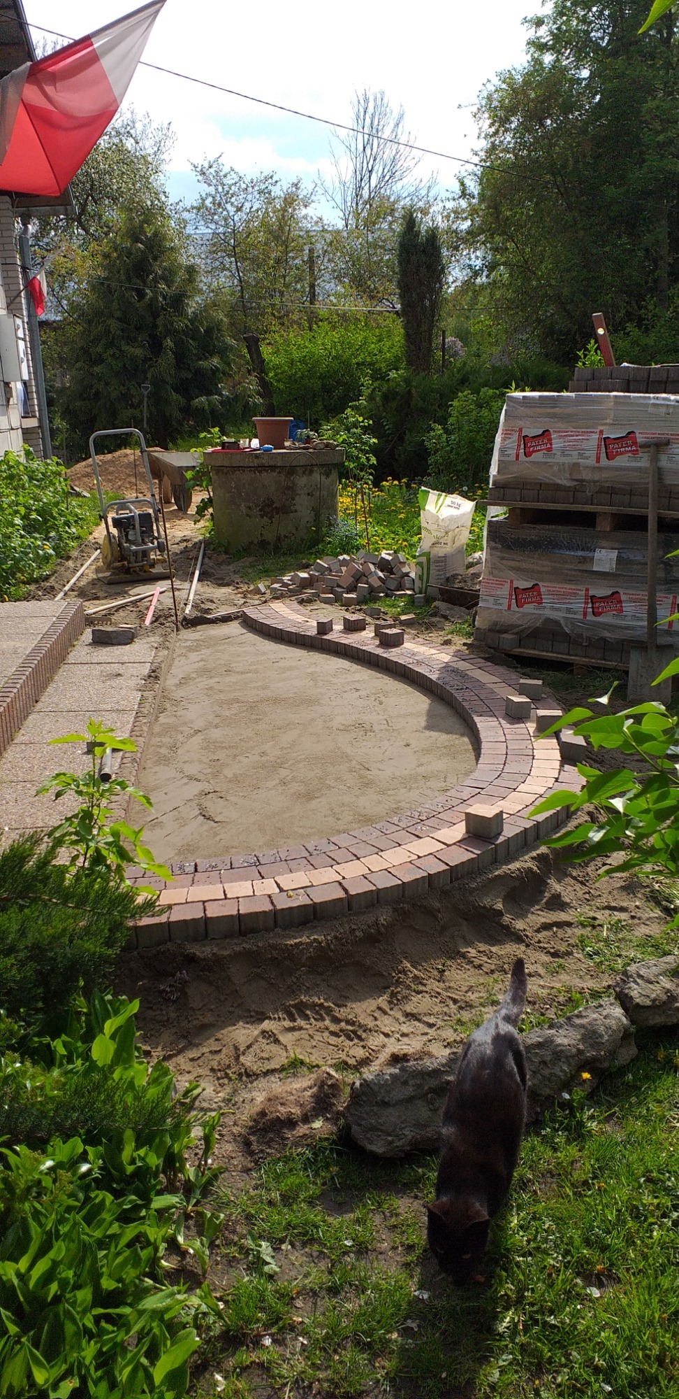 Przygotowanie podłoża pod ścieżkę z kostki brukowej w ogrodzie, widoczne obrzeża i częściowo ułożone kostki, zagęszczarka, paleta z kostką, w tle studnia, na pierwszym planie czarny kot.