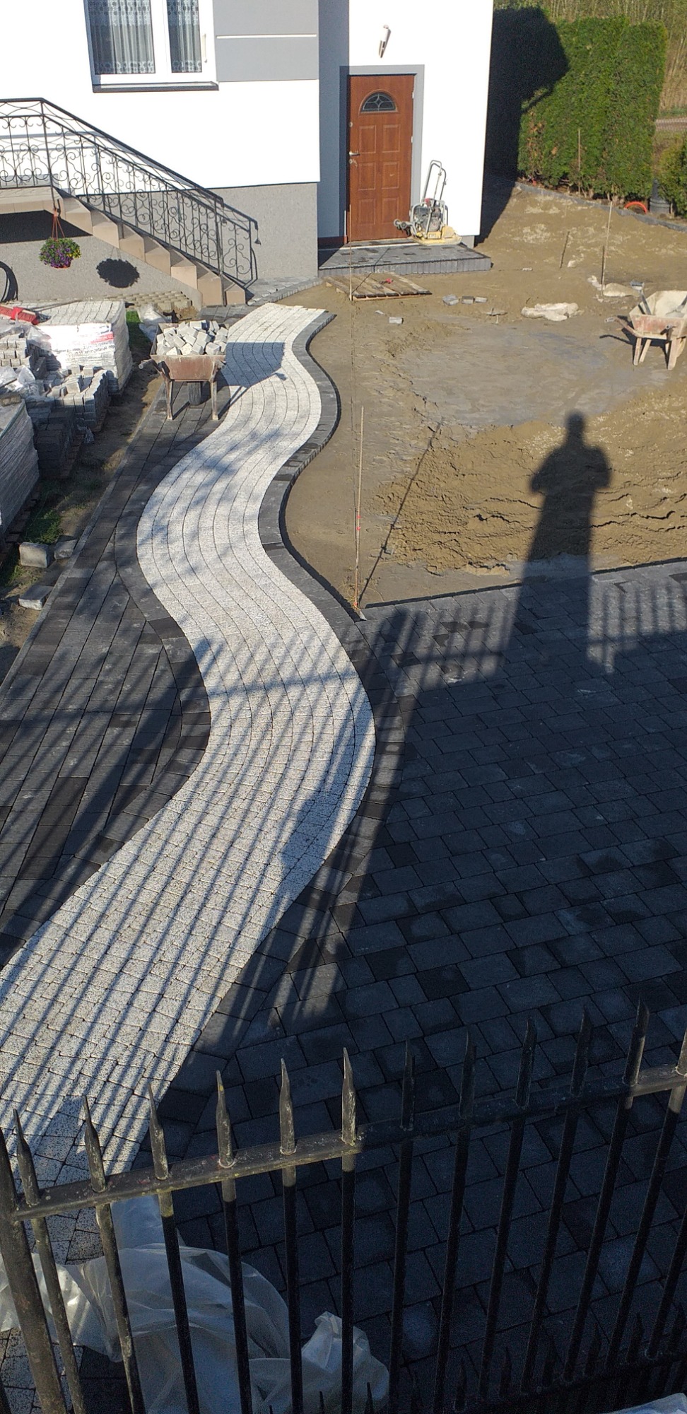 Ścieżka z jasnej kostki brukowej, wijąca się przez ciemniejszą nawierzchnię, w tle dom z drewnianymi drzwiami i cień sylwetki osoby